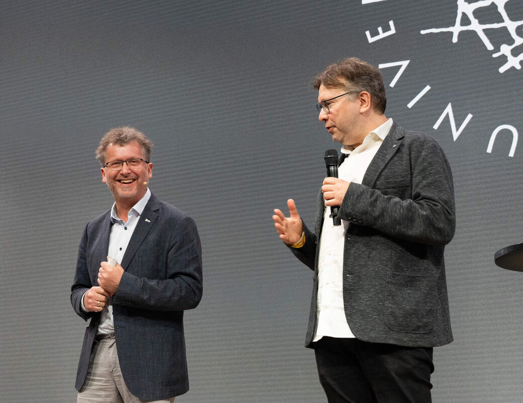 Two men in dark jackets speak on stage; one holds a microphone in front of a grey backdrop.