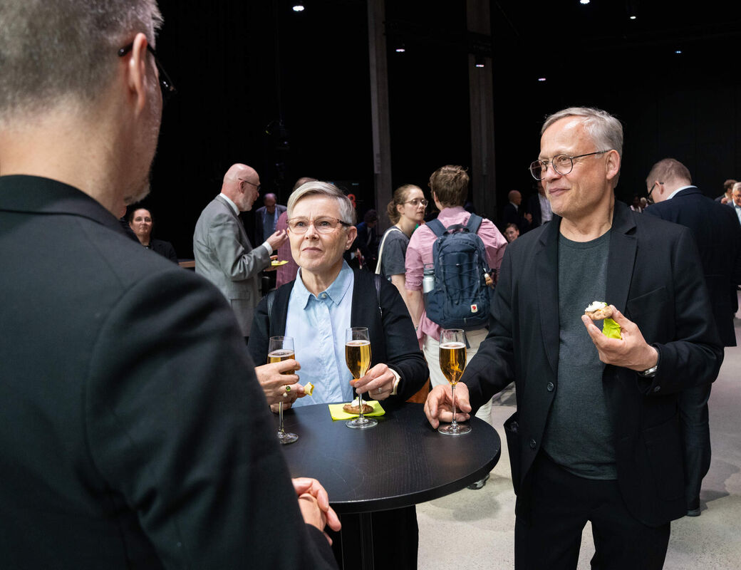 People in dark suits stand at a round table with drinks and snacks at a social event indoors