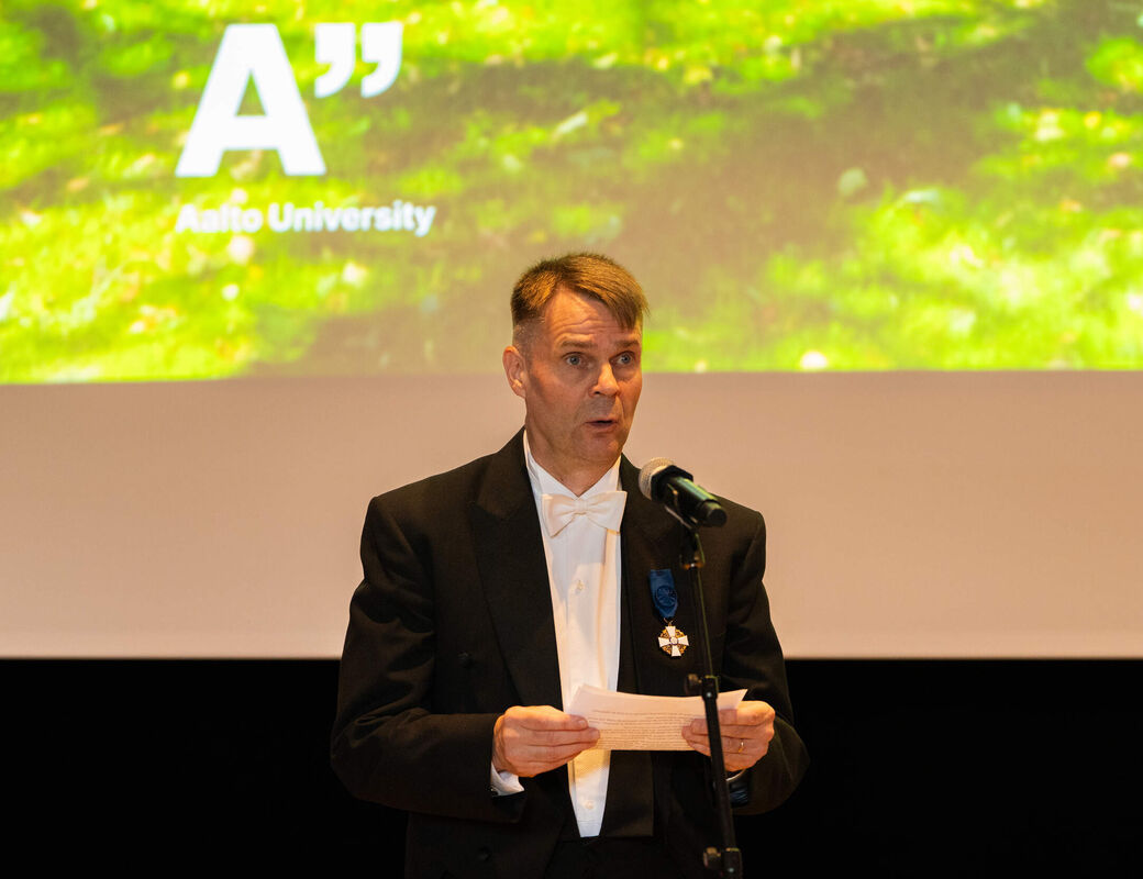 Person in black tailcoat speaking at a microphone, Aalto University logo on bright green screen behind