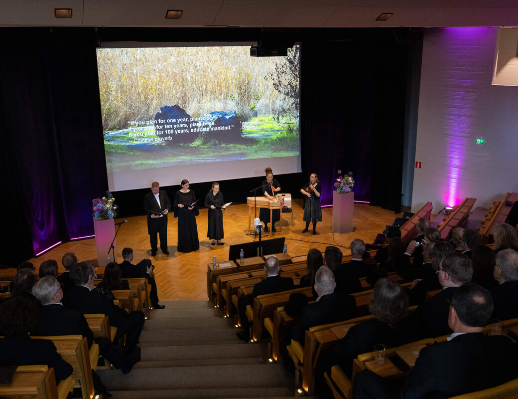Audience in a modern hall watching four speakers on stage under a large nature image with a proverb.