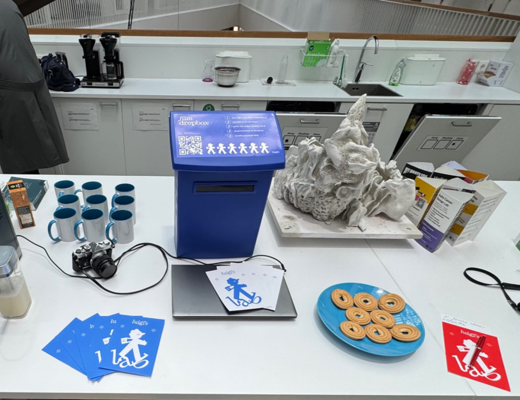 Office kitchen counter with blue film dropbox, camera, mugs, cookies on a plate and a white sculpture.