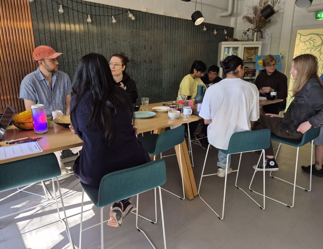 Group of people chatting around a long wooden table with drinks and snacks in a bright kitchen