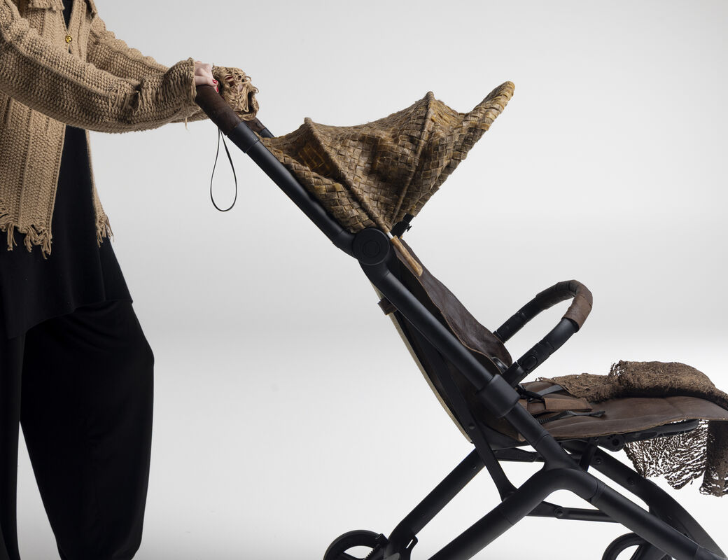 Person in loose beige jumper pushing an empty brown stroller with textured canopy and blanket