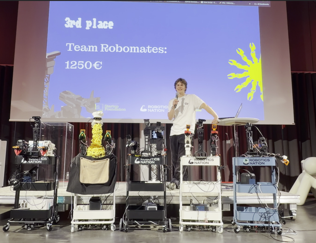 Smiling presenter with microphone stands by robots on stage under a 3rd place Team Robomates 1250€ award slide.
