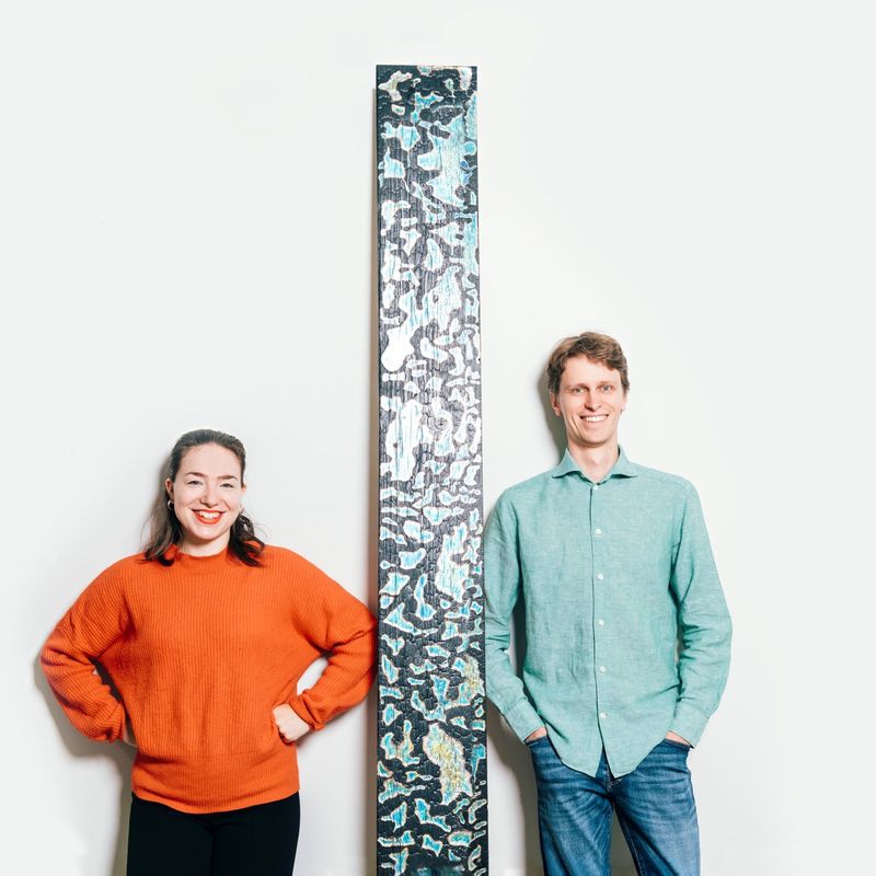 Tohtorikoulutettavat Noora Yau ja Konrad Klockars seisovat vierekkäin valkoista taustaa vasten. Heidän välissään on pystysuora puinen levy, joka on pohjaväriltään musta, mutta pinnassa on kirkkaita sinisiä ja keltaisia kuvioita. Kuva: Jaakko Kahilaniemi.
