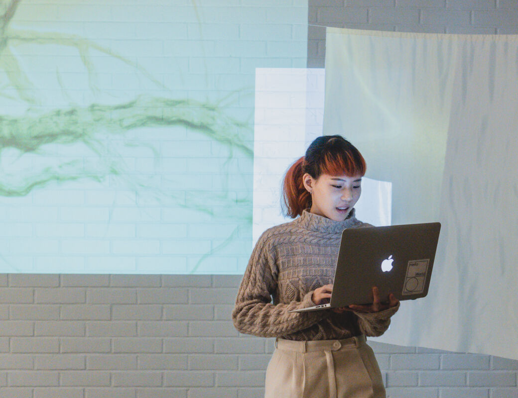 A ViCCA student stands in front of projected screens talking about her work with a macbook in hand