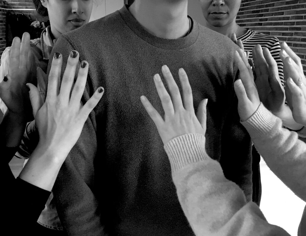 An image from the workshop Situated Knowledges and Embodied Practice. Participants stand in a circle with their hand raised, open, close, to an individual's chest. Greyscale image