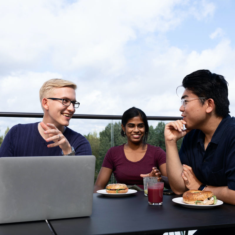 Three students eating a snack and discussing over a computer on a rooftop terrace. 