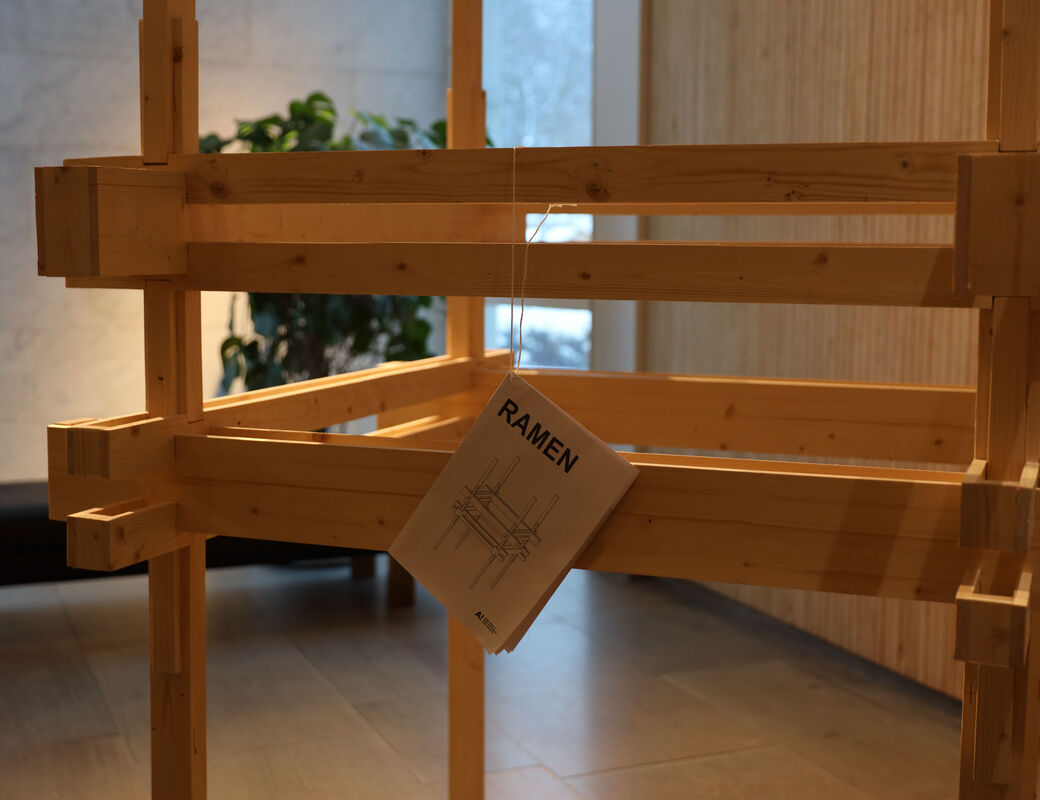 A square wooden frame with a booklet in the style of an IKEA instruction paper, hanging by a piece of string.
