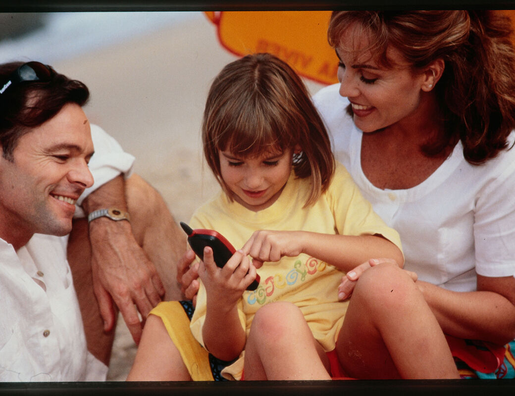 A child in a yellow shirt is using a mobile phone with two adults sitting beside them on a beach.