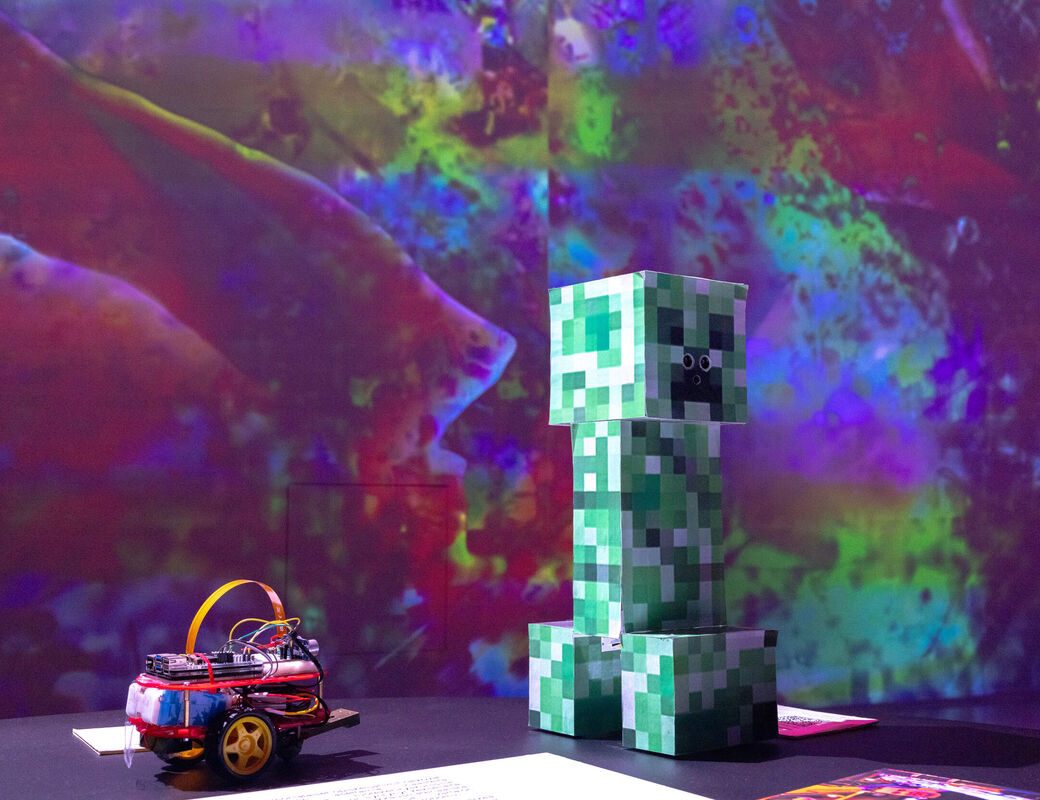 A Minecraft Creeper figure stands next to a small wheeled robot with a vivid, colourful background.