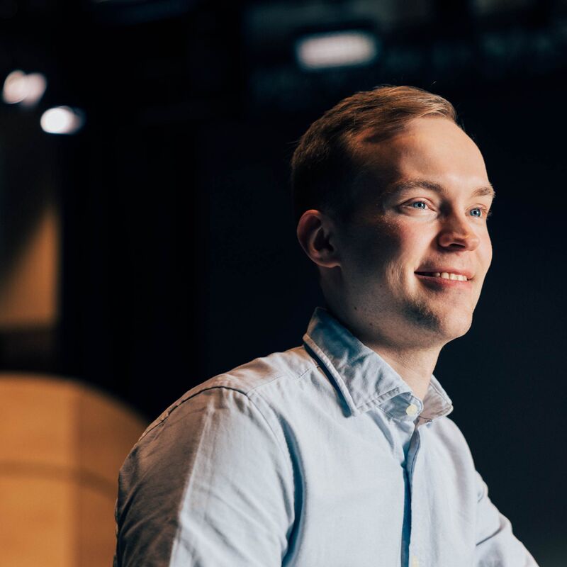 Henkilö vaaleansinisessä paidassa seisoo sisätiloissa tummalla taustalla, yläpuolella on valoja.