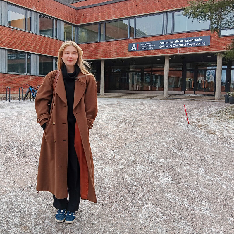 Henkilö pitkässä ruskeassa takissa seisoo rakennuksen edessä, jossa on 'Kemian tekniikan korkeakoulu' -kyltti.