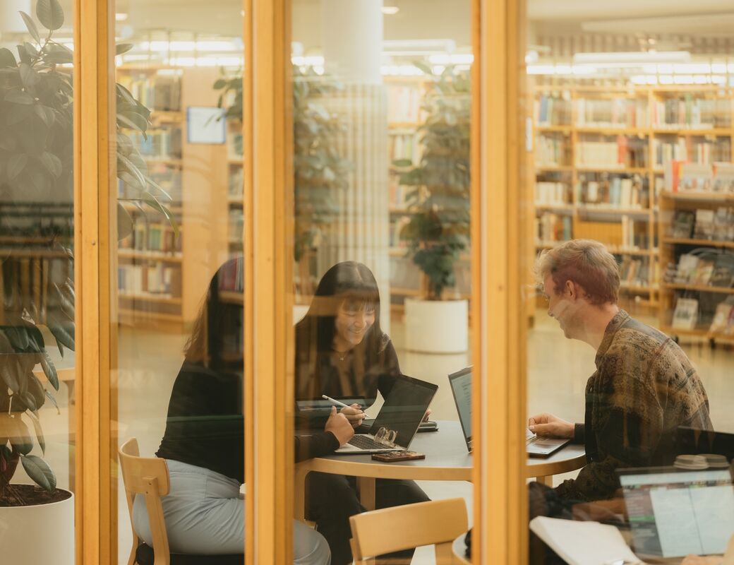 Three people working on laptops at a round table in a library, seen through glass walls.