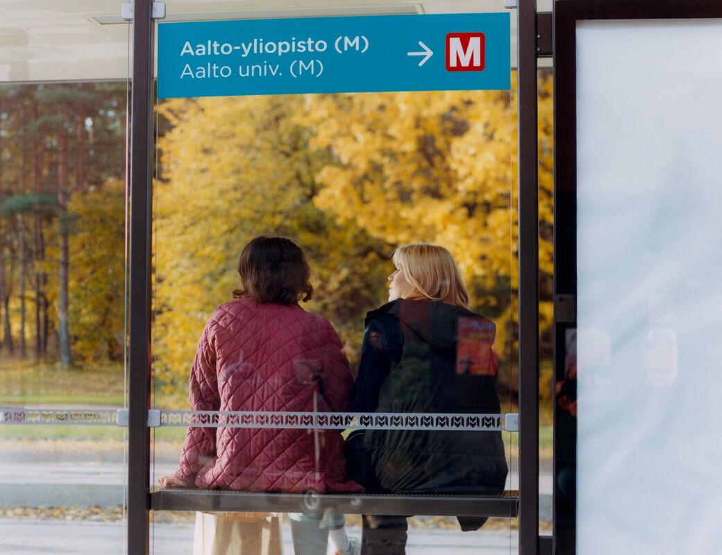 Two people sitting at a bus stop with a sign for Aalto University. Autumn trees are visible in the background.