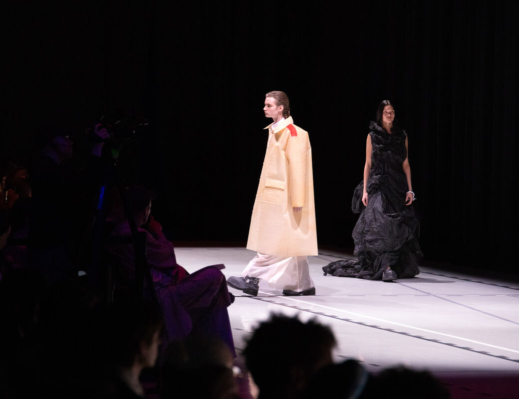 Two models walking on a runway, one wearing a beige outfit and the other in a black dress, with an audience watching.