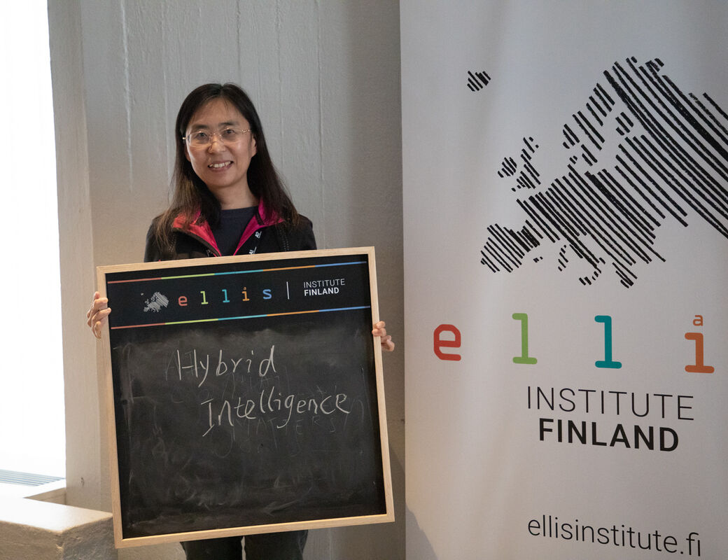 A person holds a chalkboard with 'Hybrid Intelligence' written on it in front of an ELLIS Institute Finland banner.