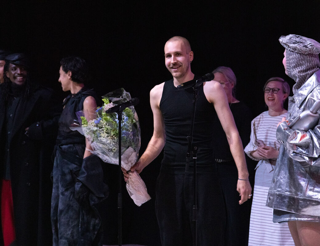 A group of people on stage, one holding a bouquet of flowers, another in a metallic costume holding a prop.