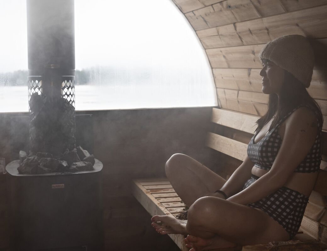A woman sits in a wooden sauna wearing a checked two-piece swimsuit and a beanie, with a stove and a misty window in the background.