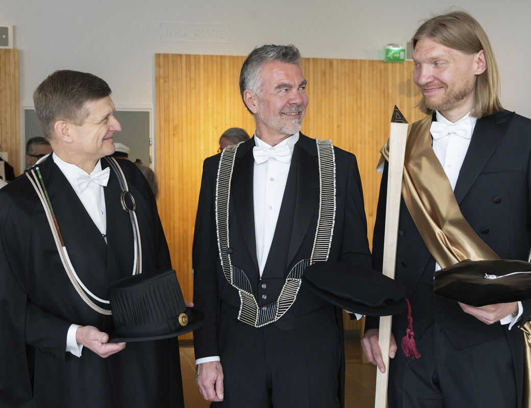 Three men in ceremonial academic garments holding formal hats and props in an indoor setting.