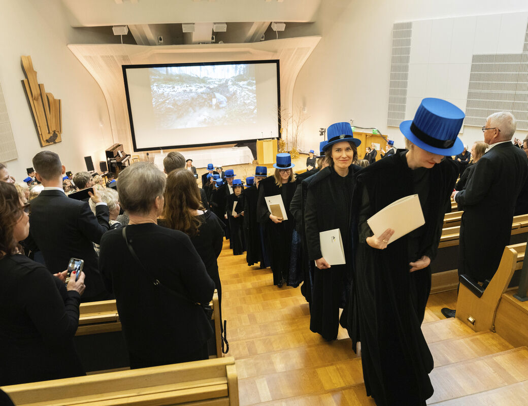 Graduation ceremony in an auditorium, graduates dressed in black gowns and blue hats holding diplomas.