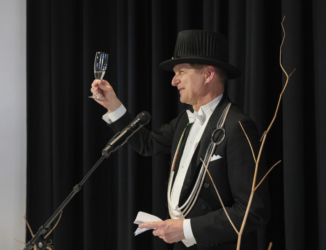 A person in formal attire and a top hat holds a champagne glass while speaking at a podium with a microphone.