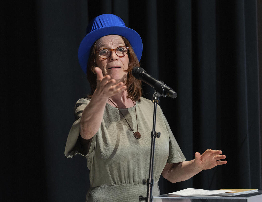 Person wearing a blue hat and grey dress speaks into a microphone on stage.