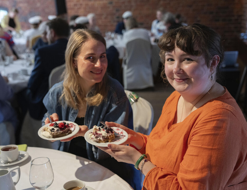 Two people holding dessert plates at a gathering. One wears an orange shirt, the other a denim jacket. Table with cups and glasses.