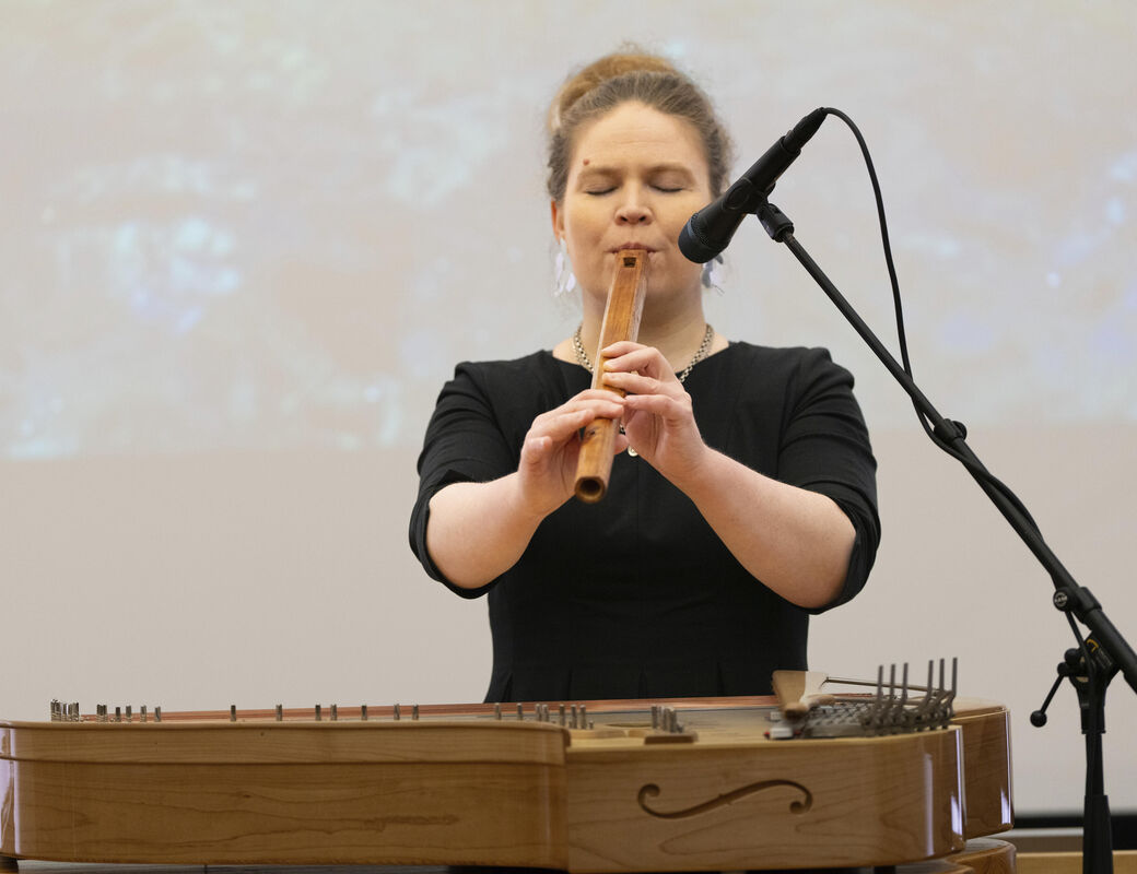 Person playing a wooden flute on stage with a zither in the foreground and a microphone stand nearby.