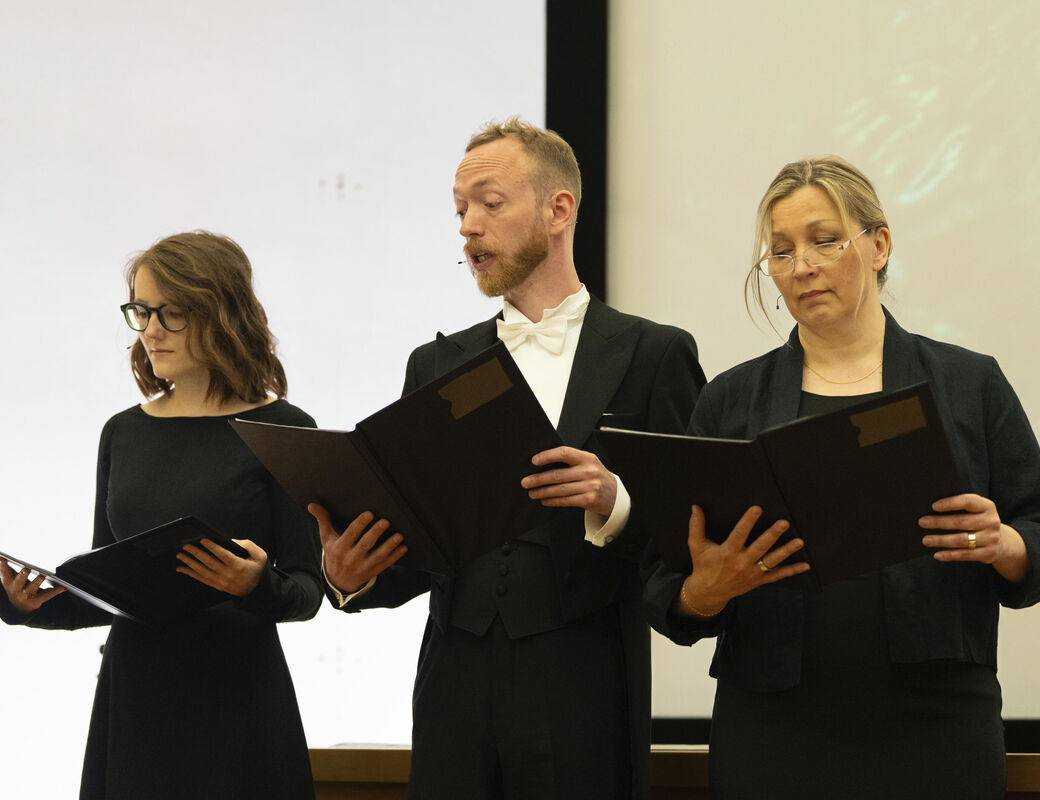 Three people dressed in formal black attire holding open black folders and appear to be singing or rehearsing.