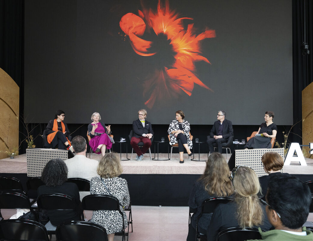 Six people sit on stage discussing, behind them a large screen showing abstract floral art. Audience watches attentively.
