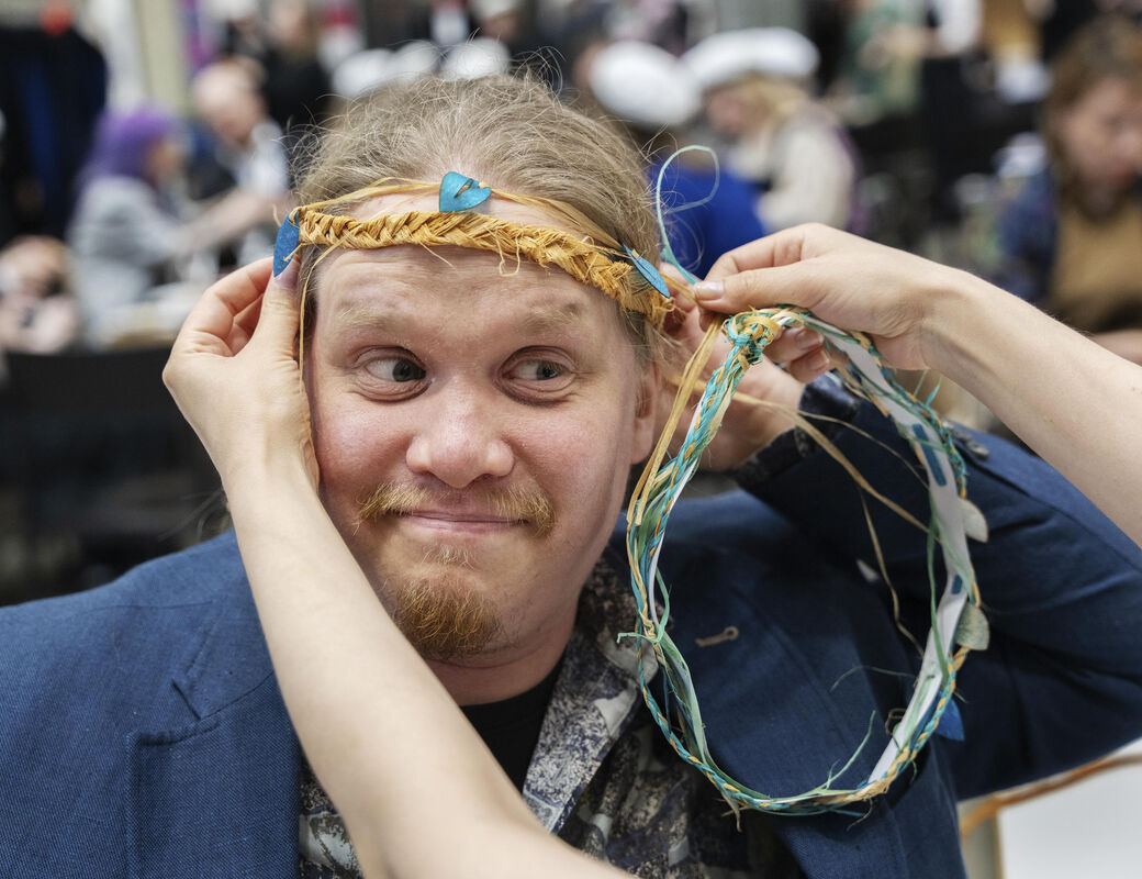 A person wearing a blue jacket is being crowned with a wreath made of colourful ribbons at a crowded indoor event.