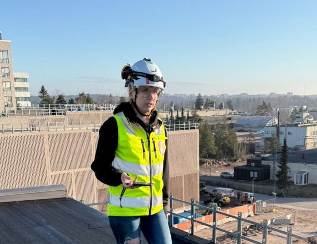Person wearing high-visibility vest and hard hat, standing on a rooftop near construction site. Blue sky.