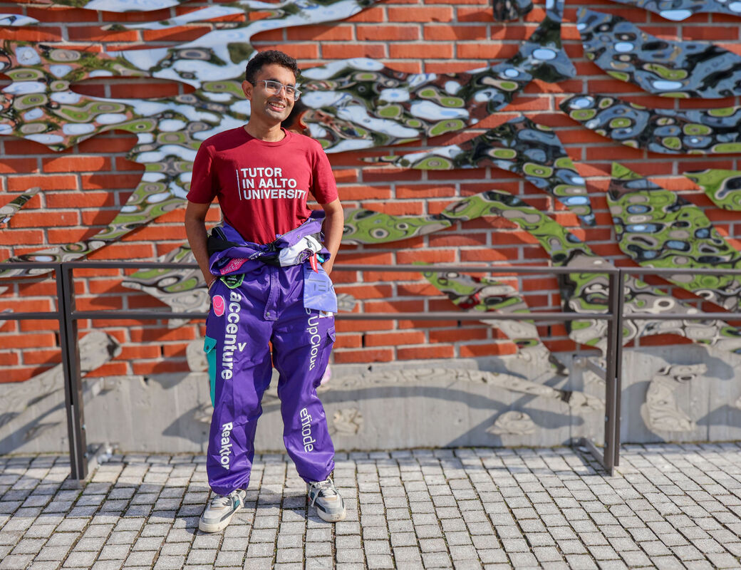 Hitesh Monga wearing Tutor in Aalto University shirt and overalls, standing in front of a brick wall with metal artwork