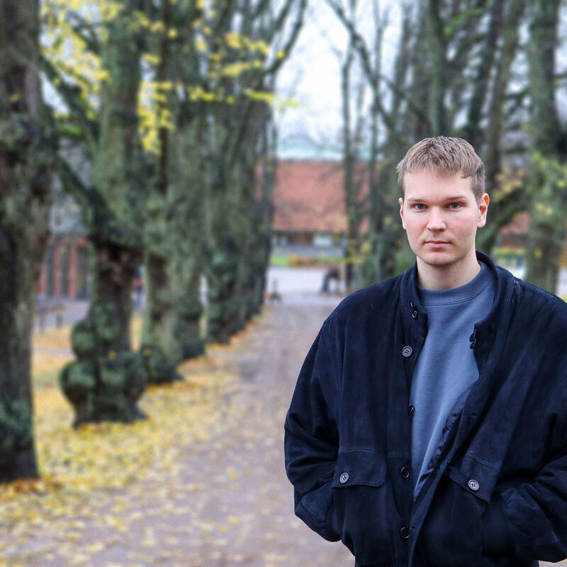 Henkilö seisoo polulla syksyisten puiden ympäröimänä, joissa on keltaisia lehtiä. Hänellä on tumma takki ja harmaa paita.