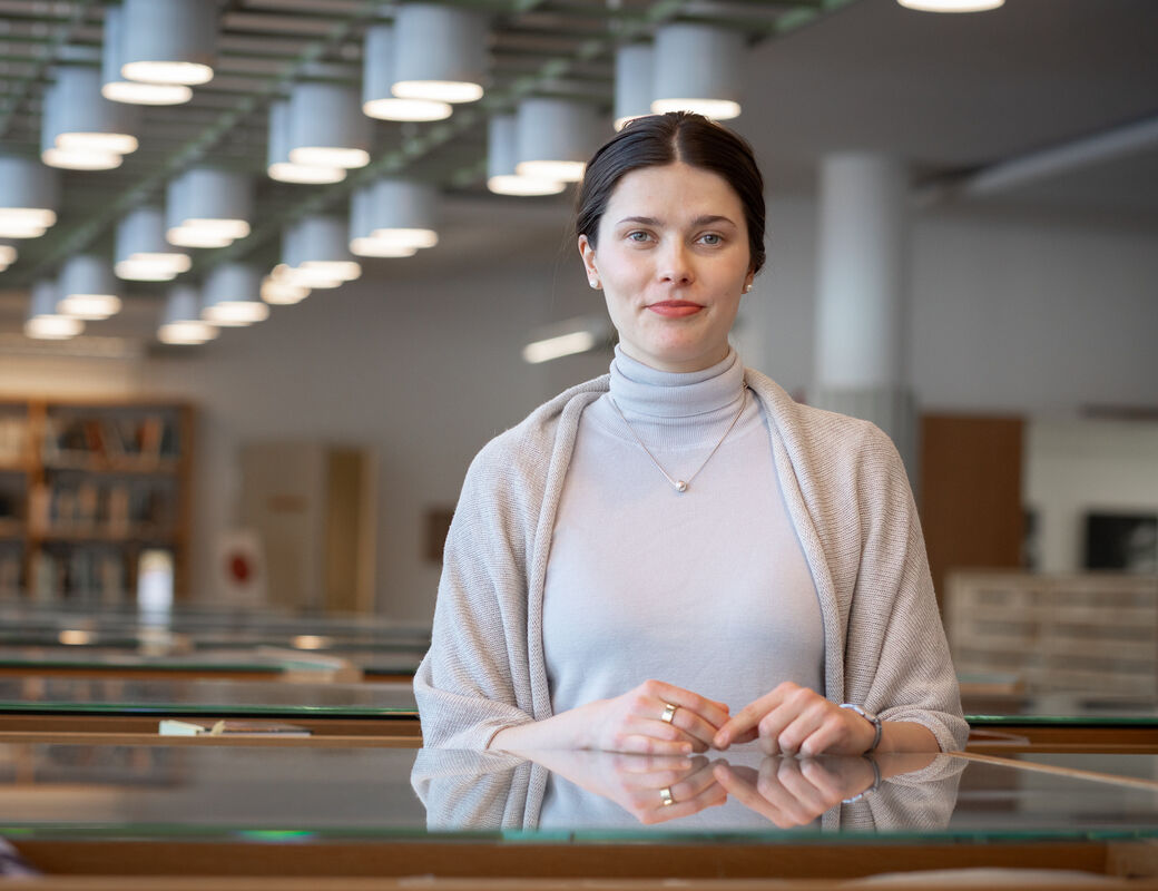 En person står i ett bibliotek, klädd i en ljusgrå polotröja och sjal. Bokhyllor och taklampor syns i bakgrunden.