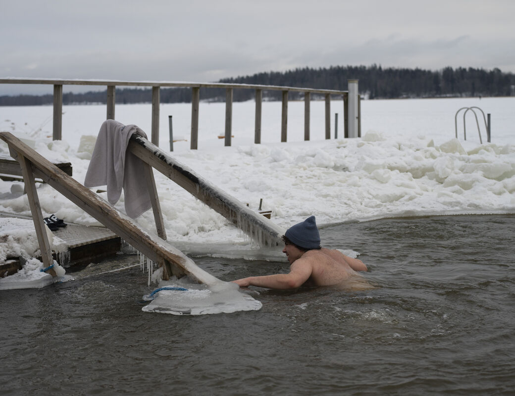 A person swims in icy water near a snowy dock with a towel hanging on a wooden railing.