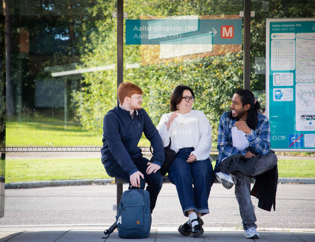 Kolme opiskelijaa bussipysäkillä, taustalla kartta ja vihreää maisemaa.