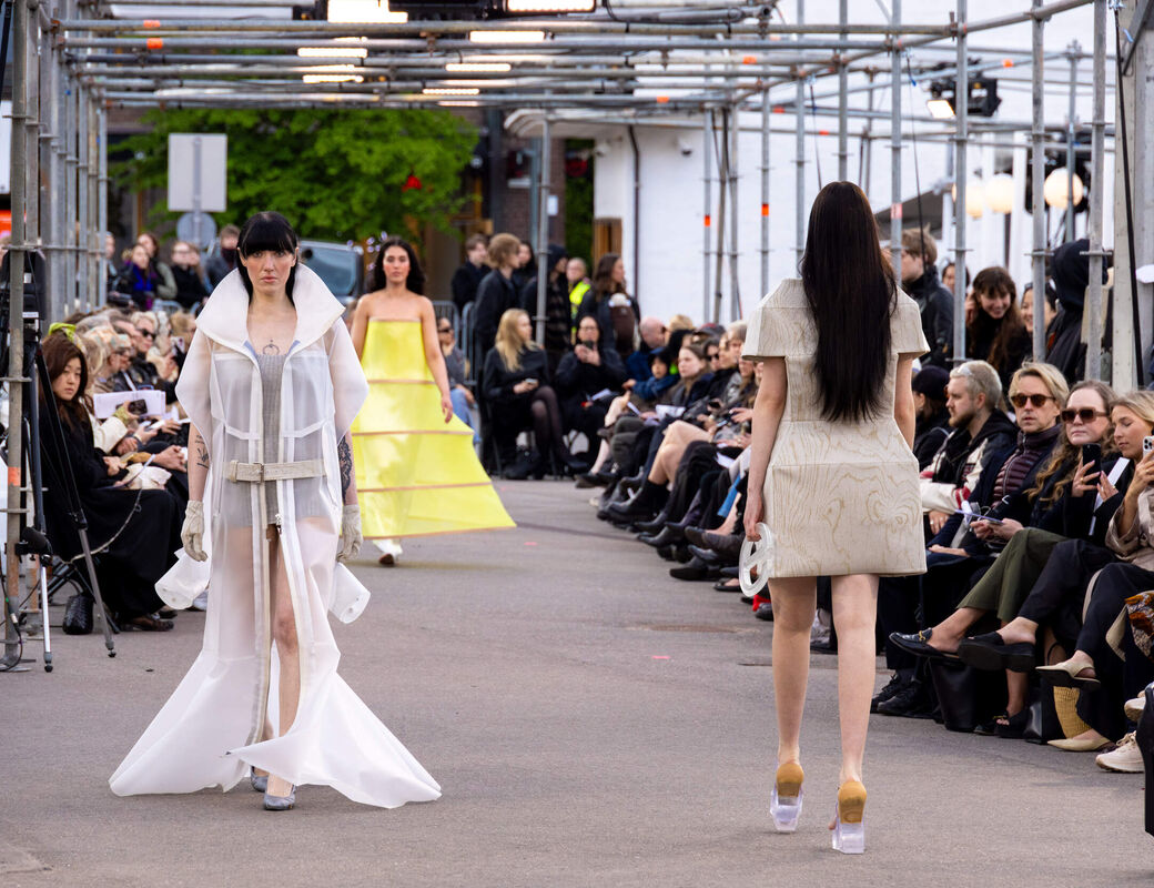 Outdoor fashion show with models in sculptural white and yellow outfits walking past seated audience