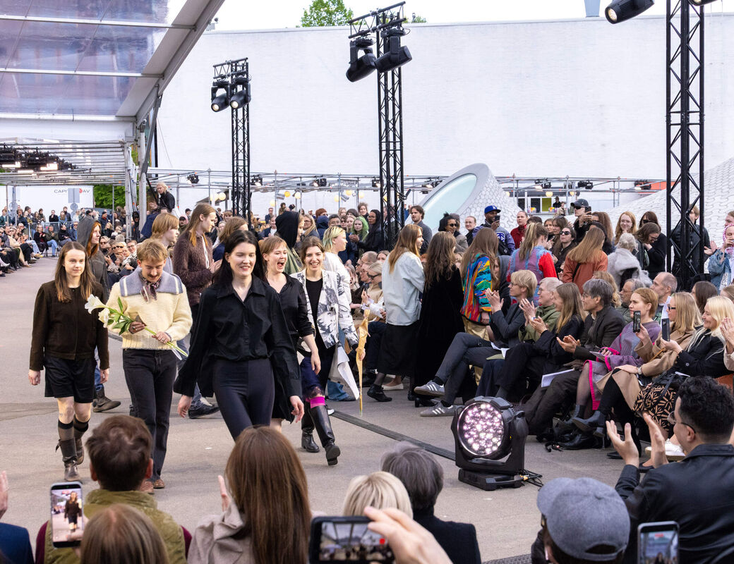 Smiling fashion designers walk on an outdoor runway, audience claps and takes photos.