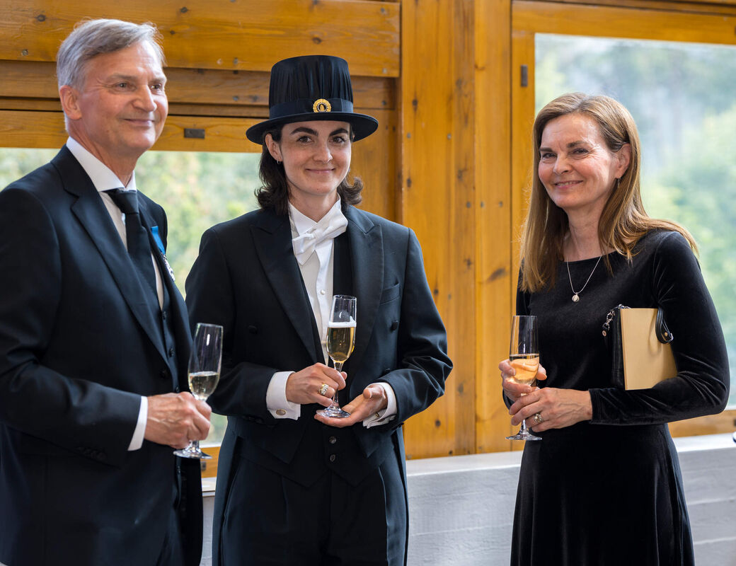 Three formally dressed people at a reception, holding drinks by large wooden-framed windows