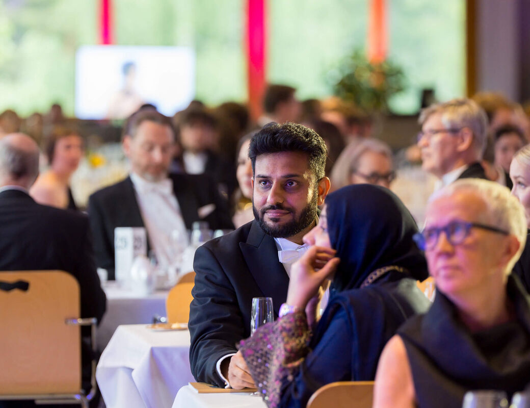 People in formal black and dark clothes seated at round tables during a bright indoor banquet