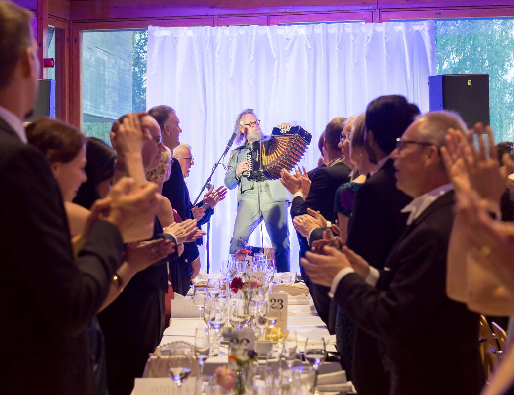 Accordionist performing on stage as formally dressed dinner guests stand and applaud at long tables