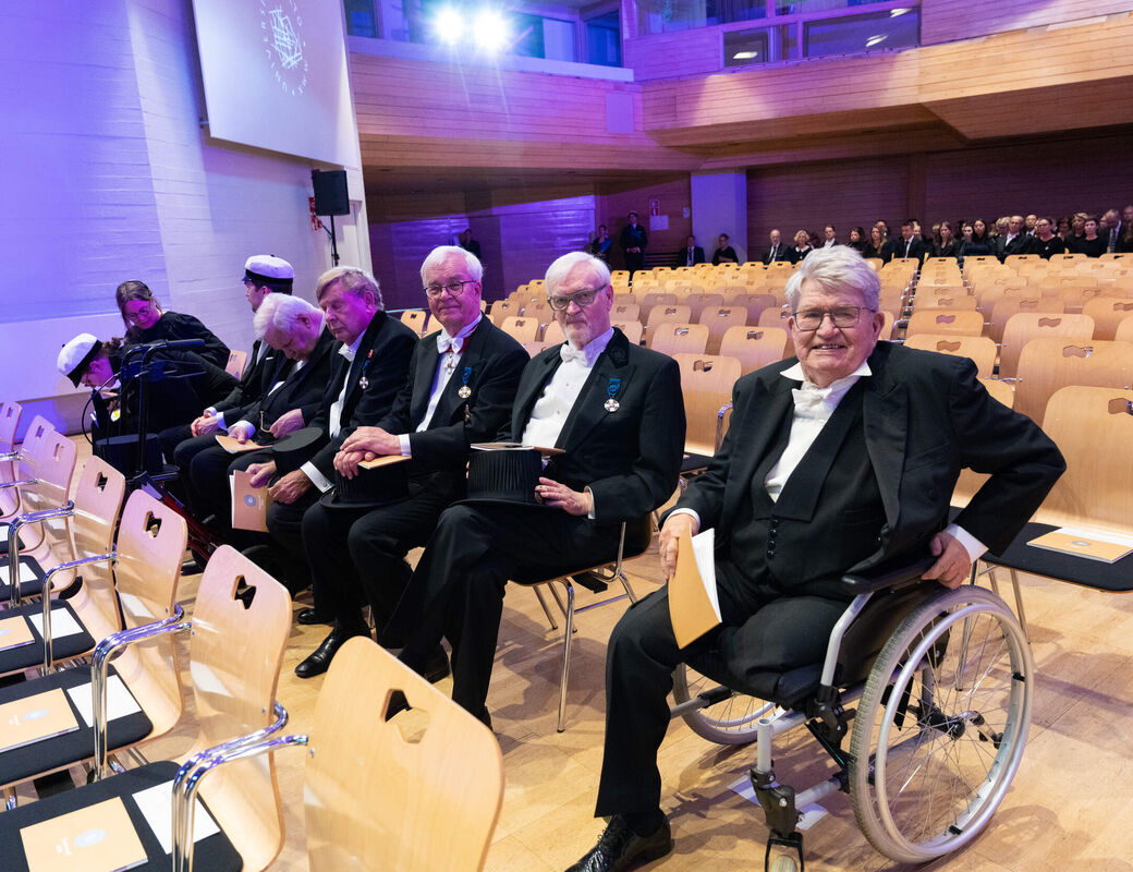 Elderly people in formal black suits sit in a bright hall, one man in a wheelchair at the front