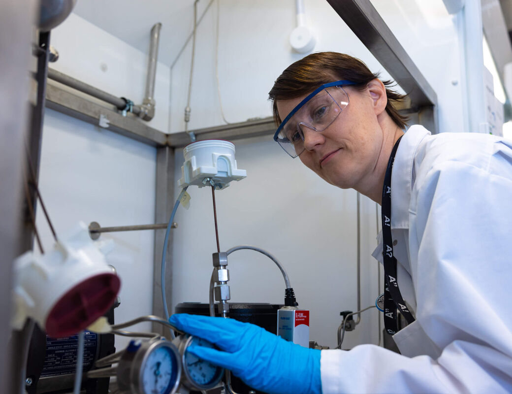 Scientist in white coat and blue gloves adjusting gauges on lab equipment in a fume hood