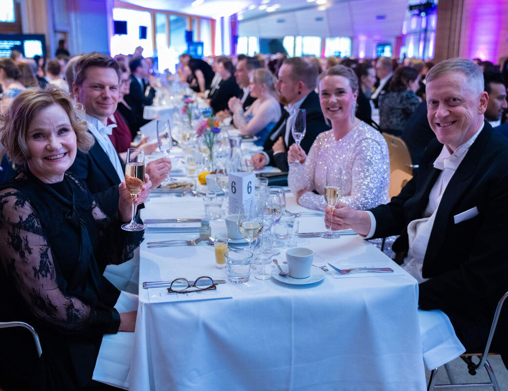 Guests in formal wear sit at a long white‑clothed banquet table, holding champagne flutes
