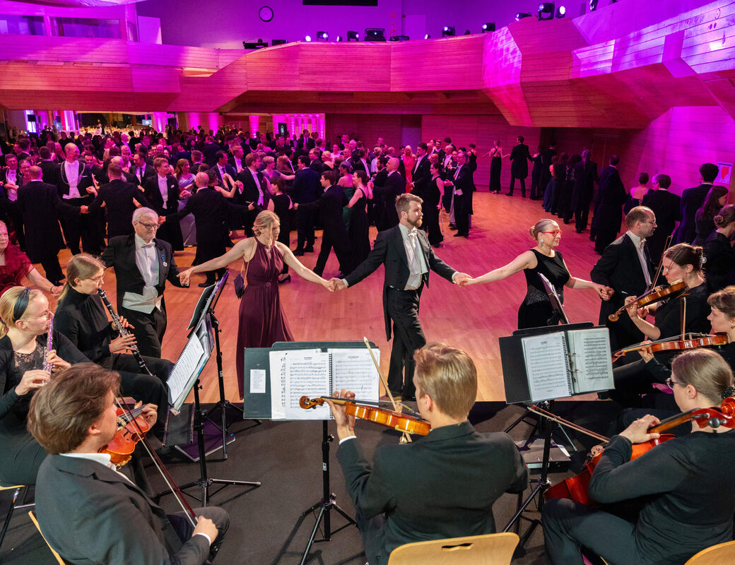 Orchestra plays on stage while couples in formal dress waltz in a bright pink-lit ballroom