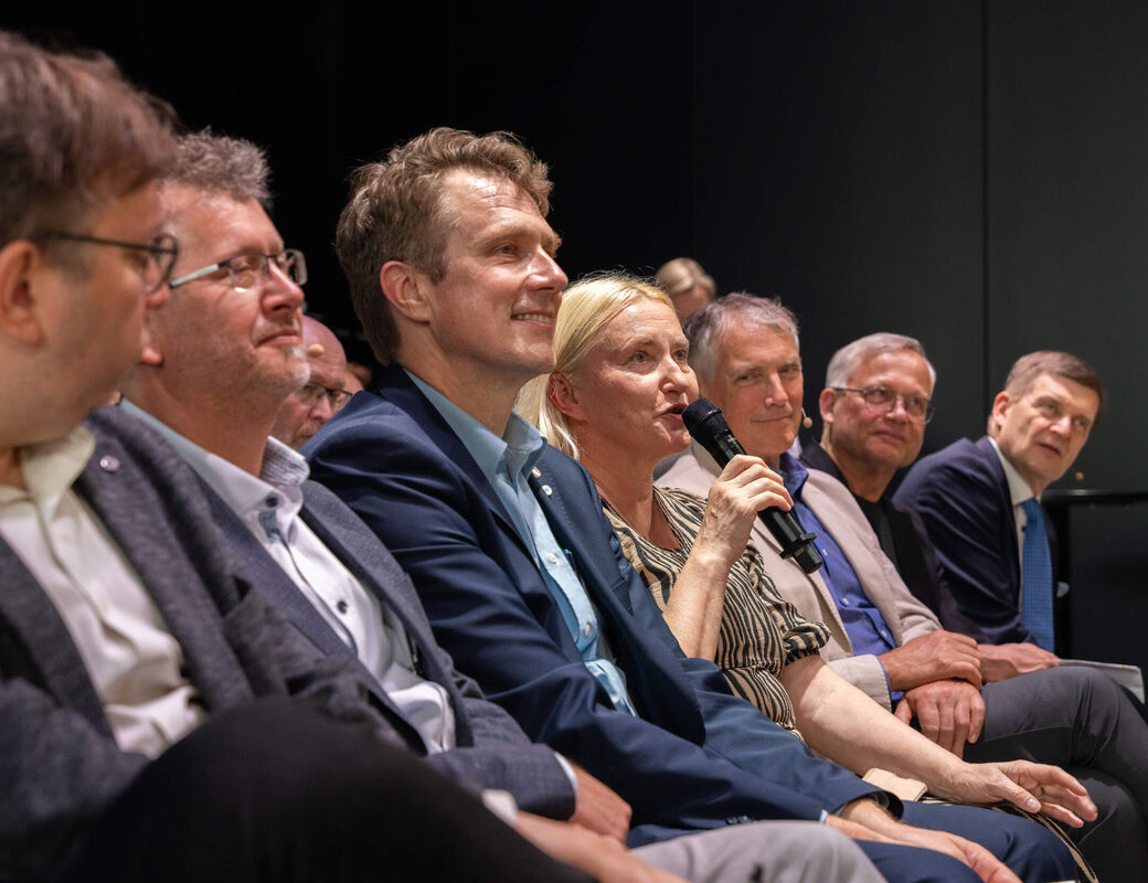 Audience row at an event, a woman with a microphone speaks while others in suits listen