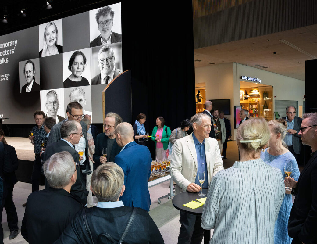 People in smart clothes networking with drinks at an indoor event, large Honorary Doctors’ Talks screen behind.