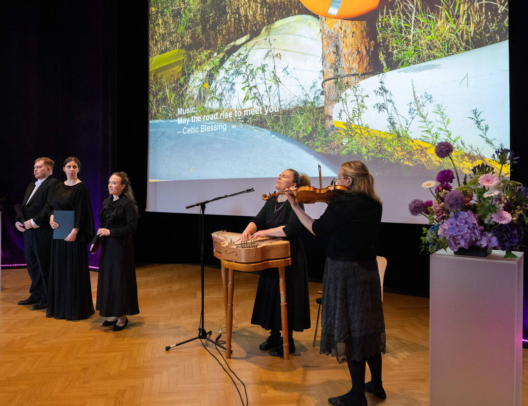 Musicians and singers perform on a wooden stage with floral displays and a large nature image projected behind.
